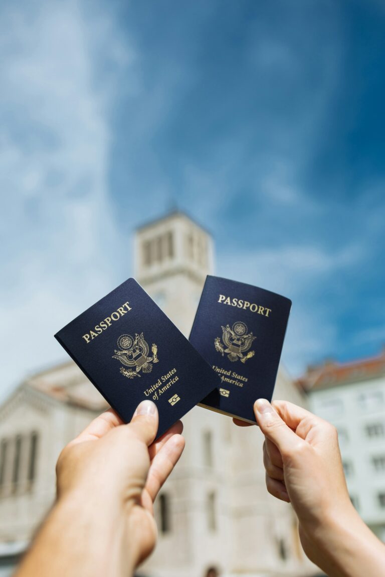 Dos manos sosteniendo pasaportes de los Estados Unidos bajo un cielo azul con un edificio desenfocado al fondo, representando servicios de ciudadanía y trámites de inmigración de USCIS.